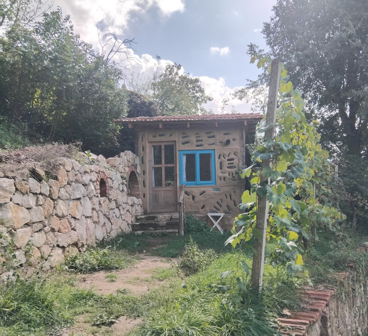 A unique cottage is framed by natural stone walls and greenery. The entrance features a wooden door with blue accents, and there are steps leading to the entry. A small white chair is positioned nearby among the lush vegetation.
