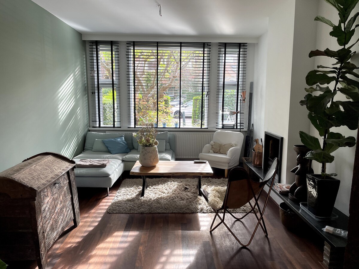 A bright living space features large windows adorned with wooden blinds, allowing natural light to fill the area. A comfortable sectional sofa and armchair are positioned around a rustic wooden coffee table. A potted plant adds a touch of greenery, complementing the warm wooden floors.
