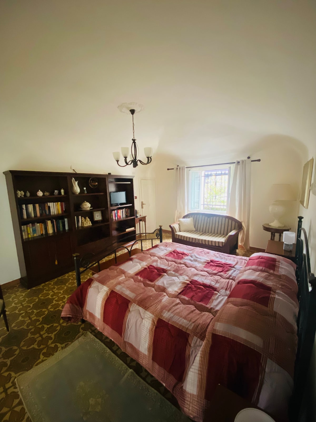 A cozy bedroom is illuminated by natural light from a nearby window, adorned with sheer curtains. A spacious bed with a patterned blanket occupies the room, accompanied by a decorative seating area and a dark wooden bookshelf filled with books. A chandelier adds to the room's ambiance.