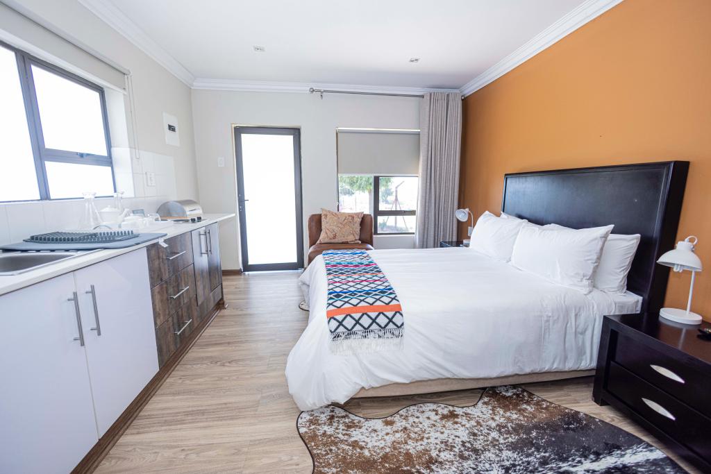 A bright and open space features a spacious bed dressed in white linens, complemented by a colorful throw. A kitchenette is visible in the background, with large windows allowing natural light and a cozy armchair near the entrance.