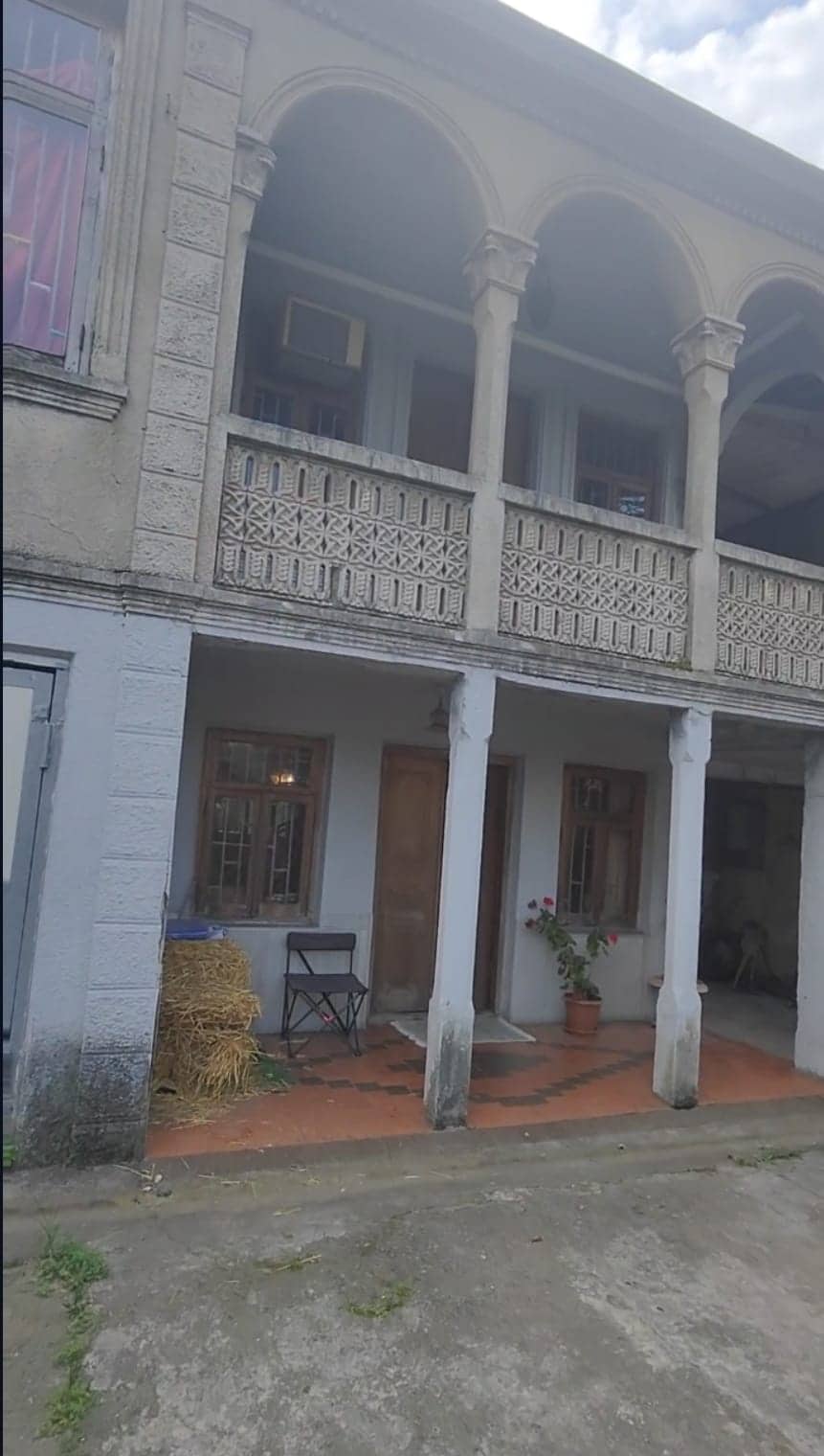 An exterior view of a two-story building is presented, featuring decorative archways and a balcony with intricate wooden detailing. The ground level includes an entrance flanked by windows, a potted plant, and a small patio area with a chair. Natural light highlights the textured surfaces.