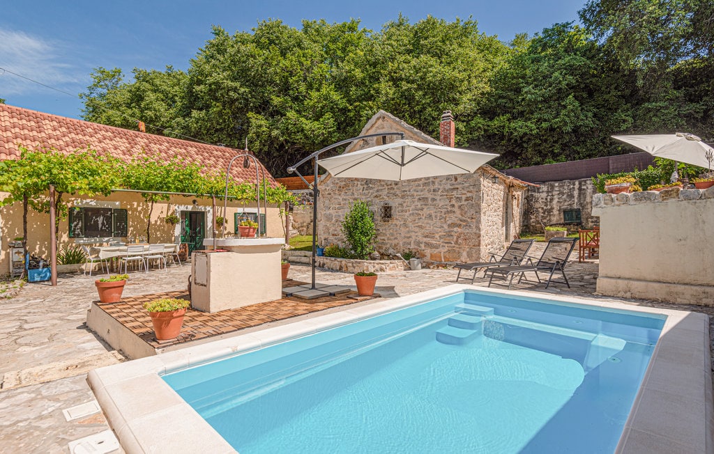 A refreshing pool is framed by a stone patio, with lounge chairs positioned nearby under shaded umbrellas. Lush greenery surrounds the area, while two charming houses can be seen in the background, creating a serene environment for relaxation and leisure.