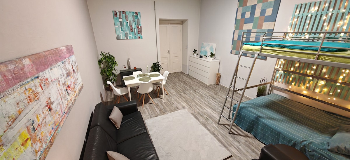 An open living space features a sleek leather sofa facing a large art piece. A dining area with a round table for four is visible, complemented by a dresser and plants. A bunk bed occupies one corner, adorned with colorful bedding and soft lighting.