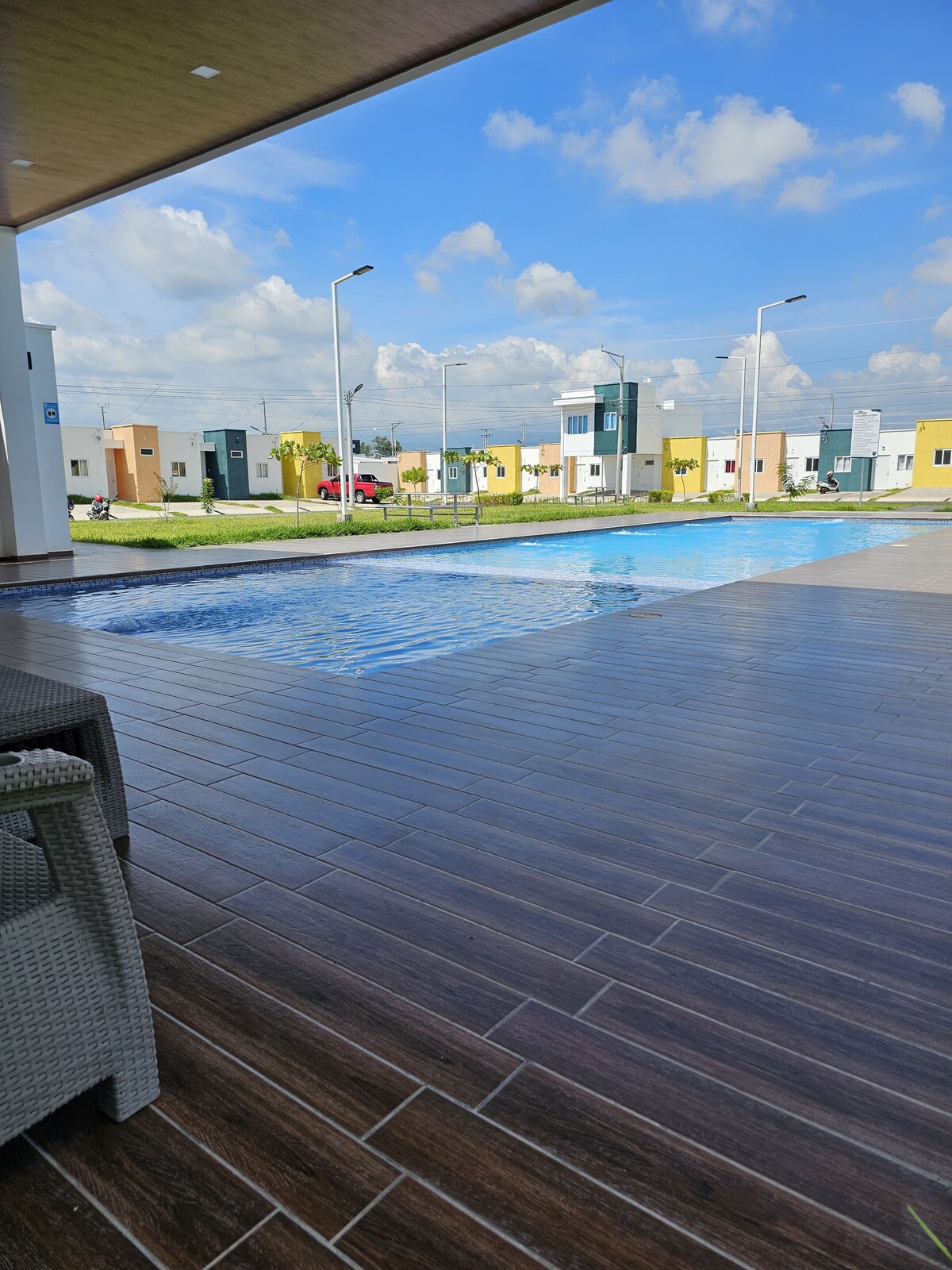 A clear view of a tranquil pool is presented, reflecting the blue sky and surrounding greenery. Comfortable seating is positioned nearby, offering a relaxing spot to enjoy the outdoor space. Modern structures are visible in the background, providing a sense of community.