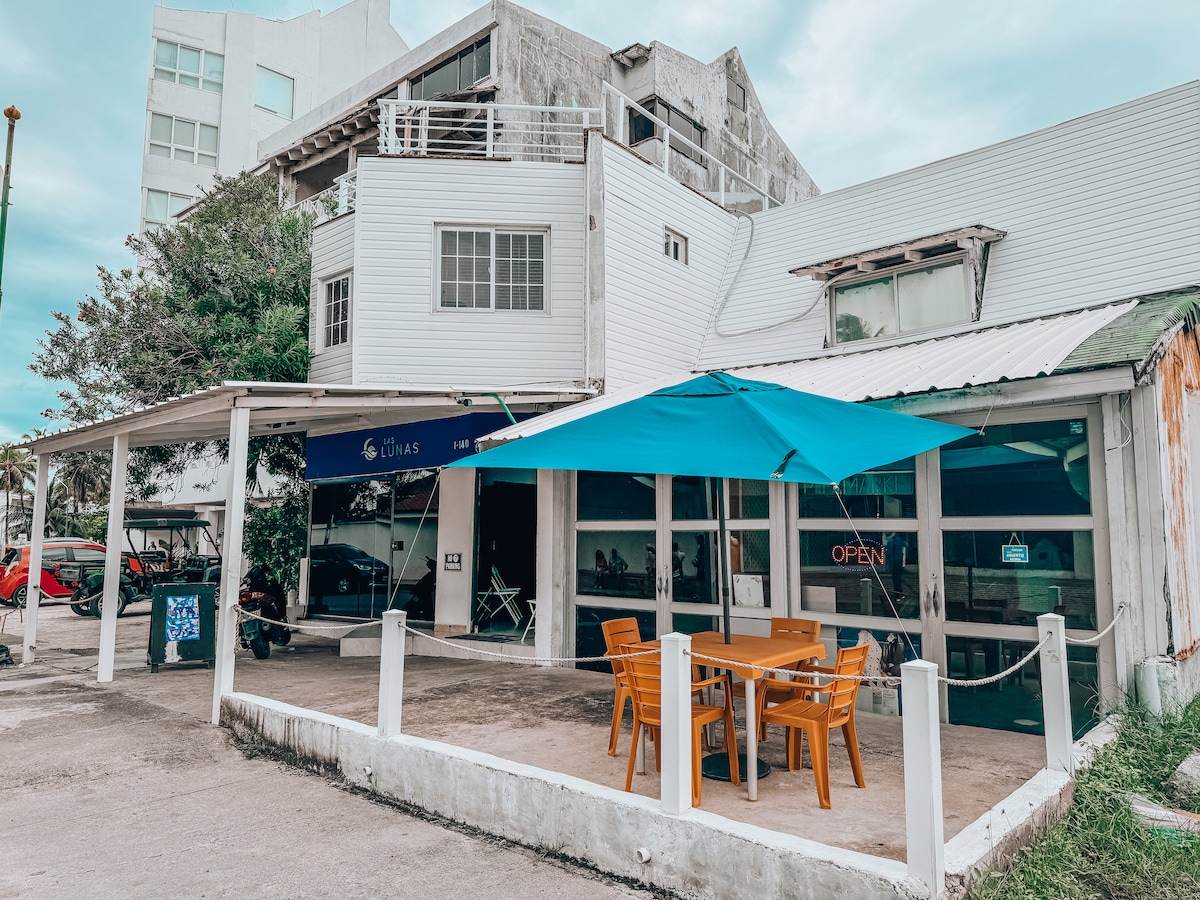 A casual café exterior is displayed, featuring bright orange furniture under a large blue umbrella. Large glass windows allow visibility into the café space, with a sign indicating 'OPEN.' Nearby, a parking area is visible, contributing to the vibrant atmosphere.