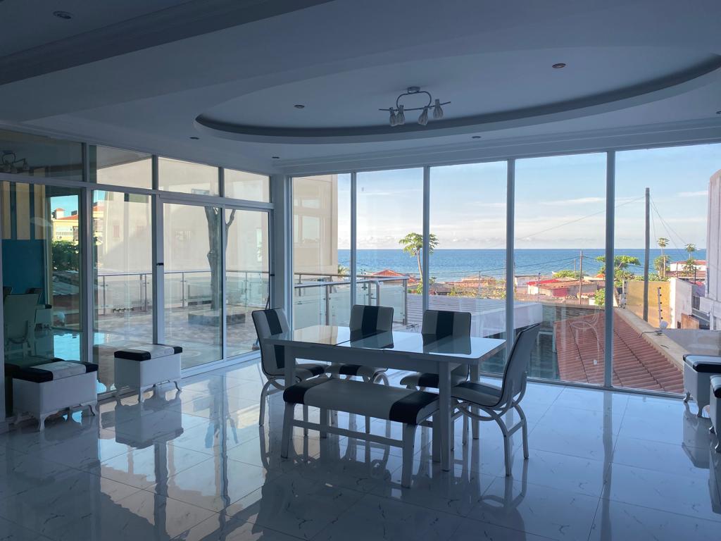 A bright dining area features a large table surrounded by elegant chairs, with expansive glass windows offering ocean views. Natural light fills the space, highlighting the polished white tiles and modern decor.