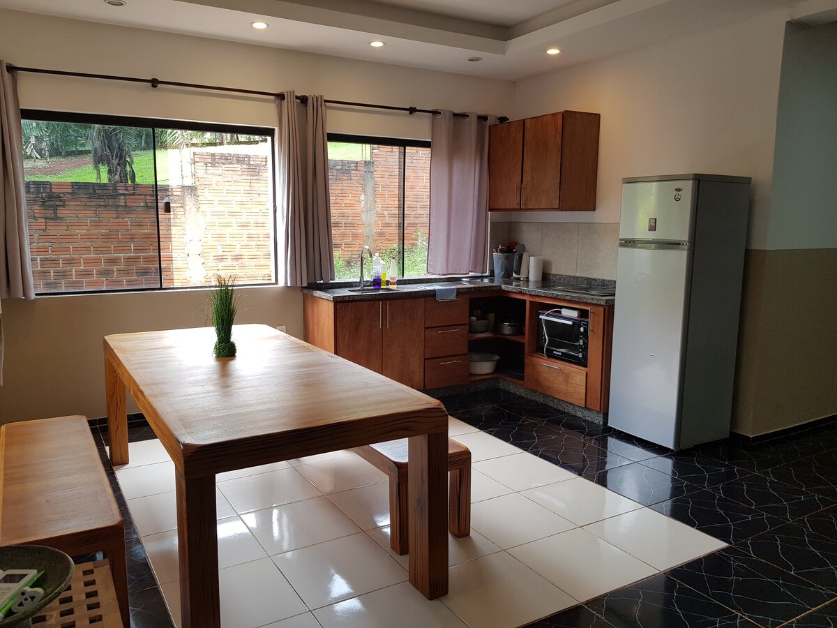 The spacious kitchen features wooden cabinetry and a large dining table, complemented by elegant seating. Natural light filters through large windows, enhancing the bright atmosphere. Contemporary appliances, including a refrigerator, are clearly visible, while plants add a touch of greenery to the space.