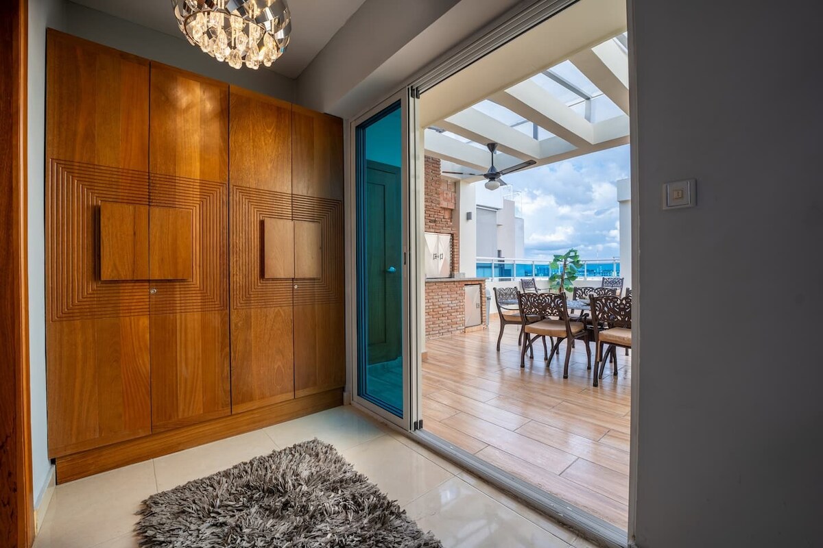 An entrance features stylish wooden cabinetry, complemented by a contemporary chandelier. A glass door leads to an outdoor seating area with a dining table surrounded by wicker chairs, providing a seamless transition to the terrace. Natural light filters through, enhancing the welcoming ambiance.