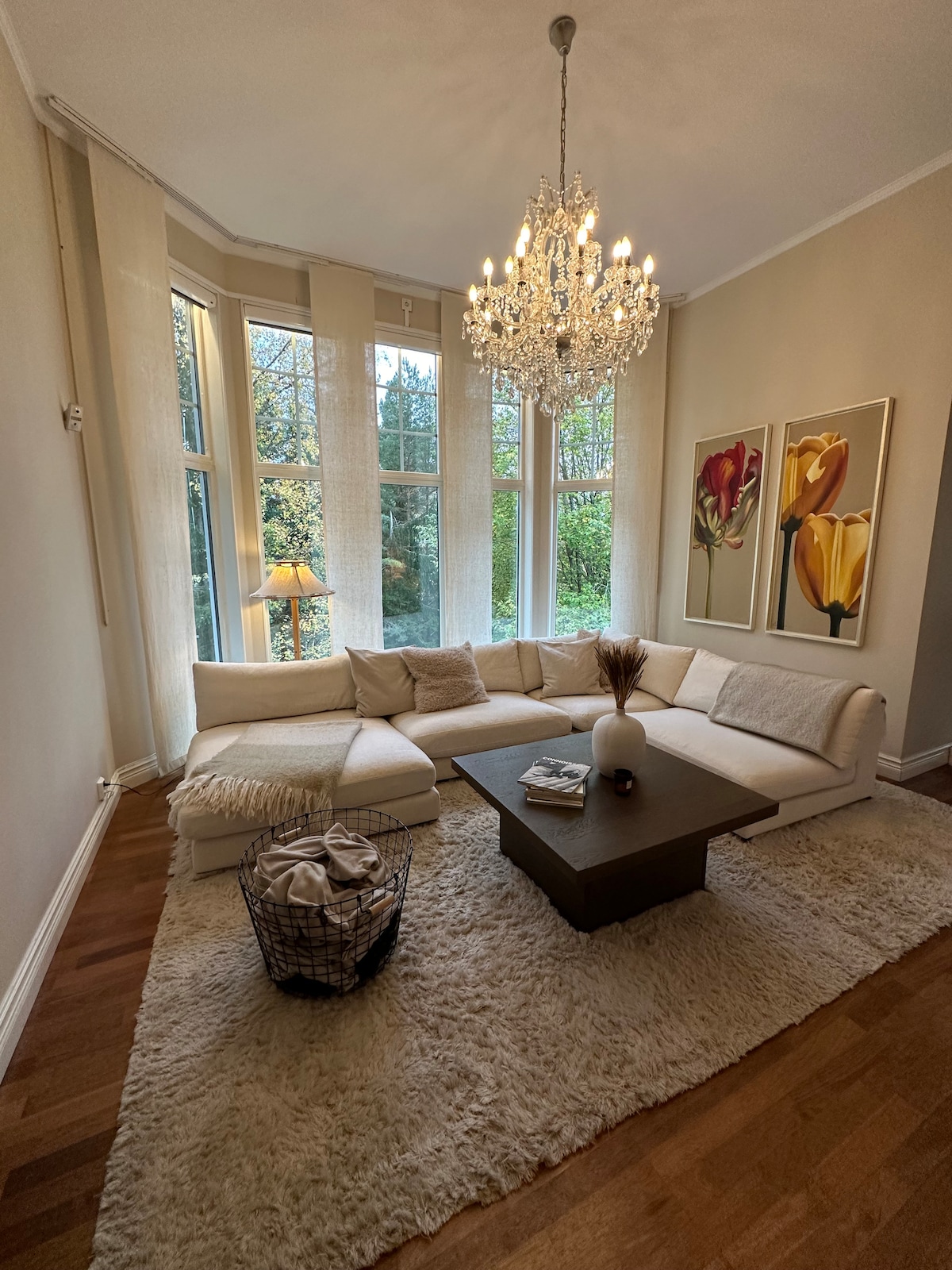 A spacious living area features a large sectional sofa adorned with soft pillows, situated on a plush rug. Natural light floods in through tall windows, complemented by elegant artwork of flowers. A decorative table sits at the center, adding to the inviting ambiance.