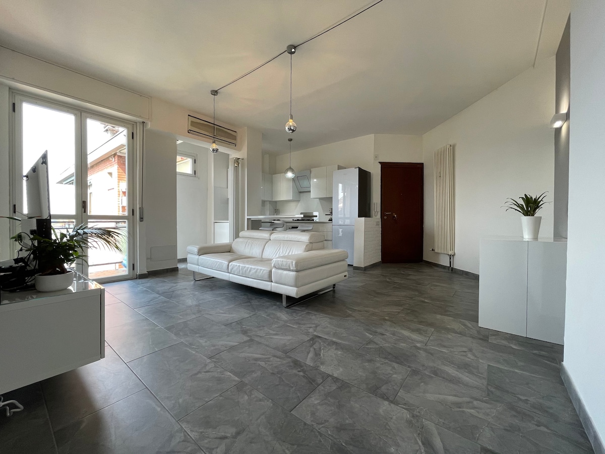 A modern living area features large windows allowing ample natural light. A comfortable white sofa is positioned in the space, complemented by sleek cabinetry and an open-plan kitchen. Minimalist decor includes a potted plant and stylish lighting fixtures, enhancing the contemporary vibe.