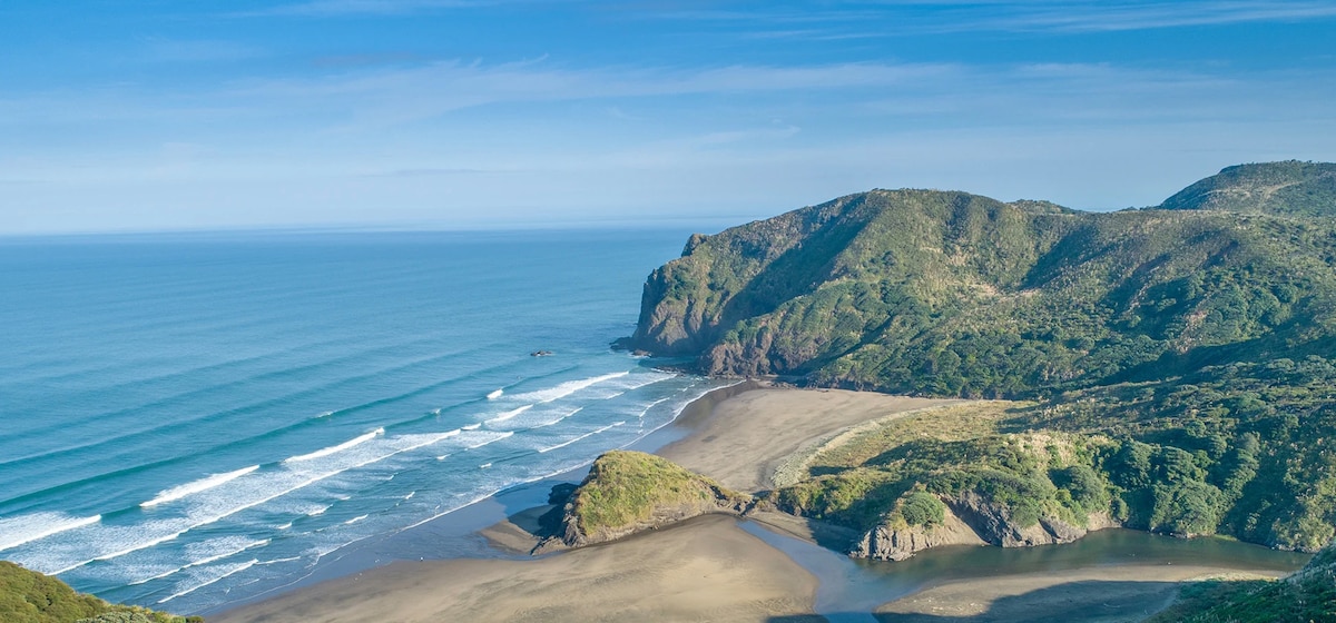 A sweeping view of a serene coastline is depicted, featuring gently rolling waves and a golden sandy beach bordered by lush green hills. The horizon meets a clear blue sky, creating a peaceful natural landscape that invites exploration.