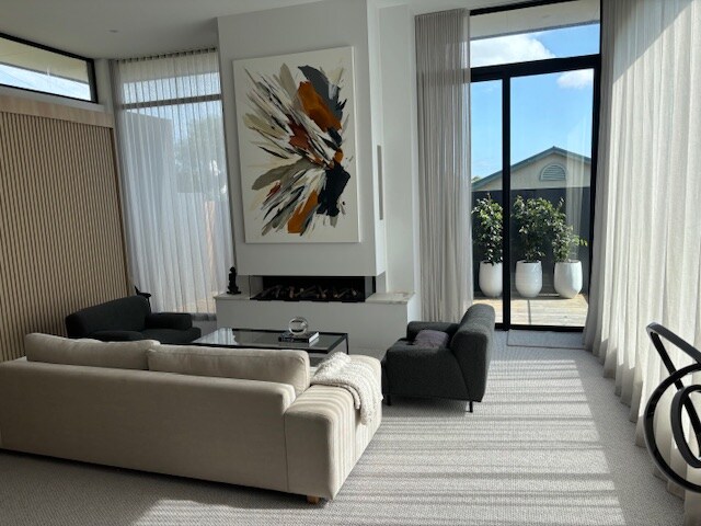 A spacious living area features large glass doors that invite natural light. A contemporary artwork piece hangs above a sleek fireplace. A comfortable sofa and accent chair are arranged around a glass coffee table, while decorative planters are visible outside through the transparent curtains.
