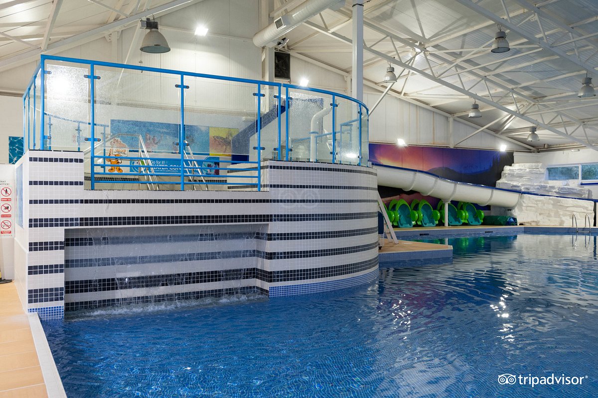 An indoor water park area features a large blue-tiled swimming pool with a gentle waterfall cascading over a smooth stone ledge. A water slide is visible, leading into the pool, while bright green slides can be seen in the background.
