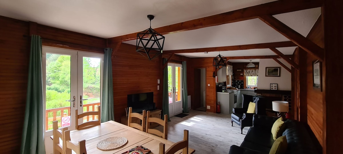 An open living area features a wooden dining table surrounded by chairs and a black leather sofa. Large windows allow natural light to fill the space, with greenery visible outside. Decorative light fixtures hang from the ceiling, adding a modern touch to the rustic design.