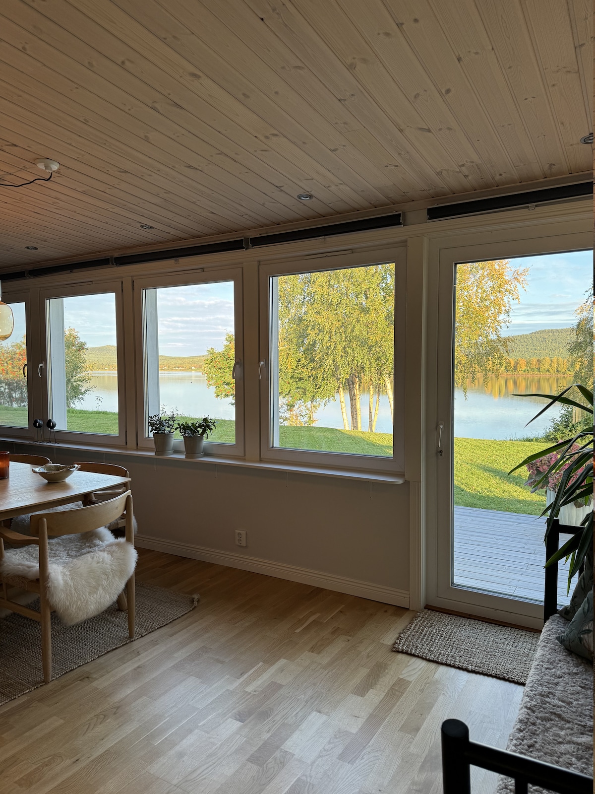 Large windows frame a scenic view of Torneälv, with lush greenery visible along the water's edge. A dining table with soft furnishings accompanies the natural light pouring into the room, creating a serene environment that emphasizes a connection with the surrounding nature.