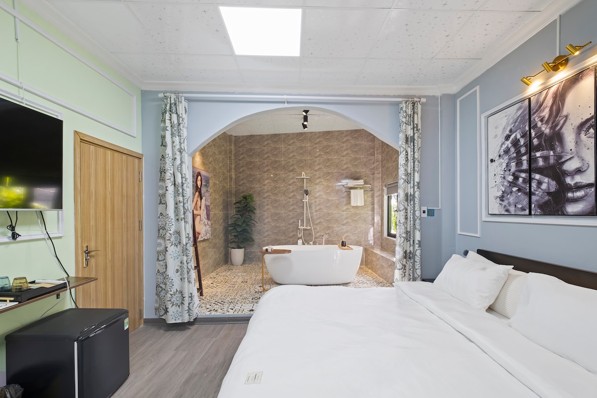 A spacious bedroom features a large bed with crisp white linens. Natural light enters through curtains, illuminating the area. A stylish bathtub is visible in an open bathroom, set against elegant mosaic tiles and greenery.