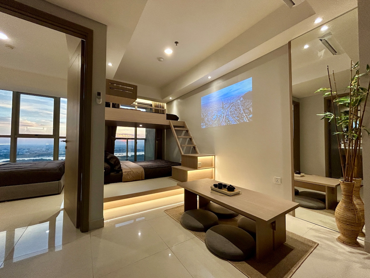 A modern living space is highlighted by a sleek projector displaying a scenic view on one wall. The room features a cozy area with a low wooden table and round cushions, complemented by a spacious bed with layered bedding, and large windows offering stunning sunset views.