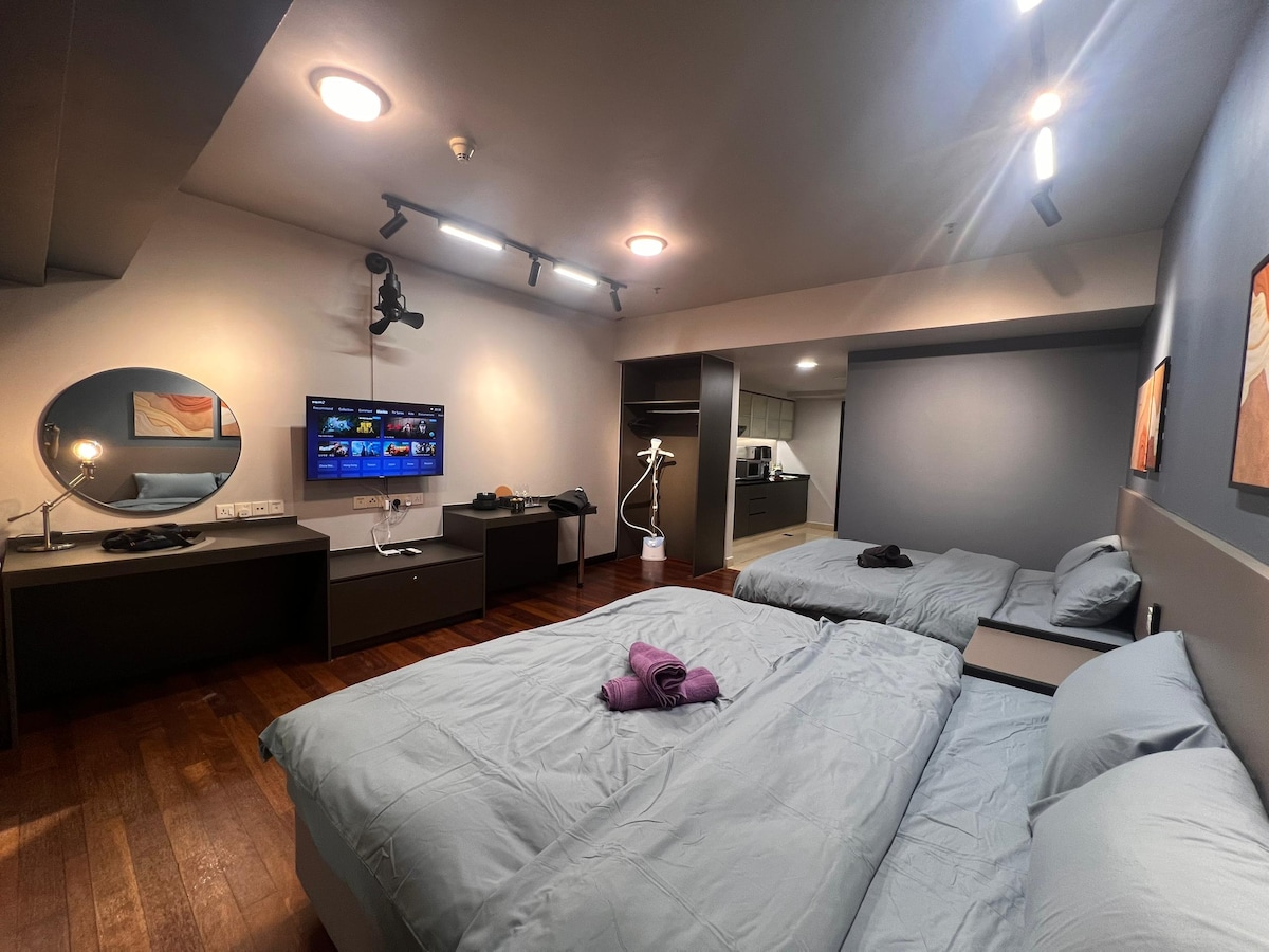 A well-lit room features two spacious beds with light gray bedding, complemented by a purple towel at the foot of one bed. A flat-screen TV is mounted on the wall, while a round mirror and a small desk with a lamp provide functional space. Wooden flooring adds warmth to the room.