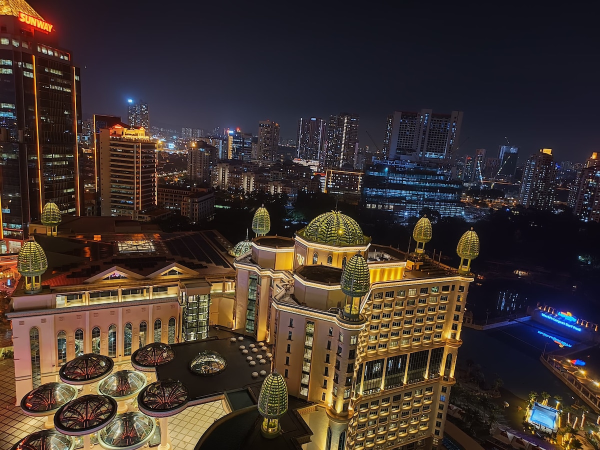 A night view showcases the vibrant city skyline, with illuminated buildings and distant lights creating a lively atmosphere. The architecture of Sunway Resort Suites, characterized by its decorative domes and balconies, is highlighted in the foreground, complementing the urban landscape.