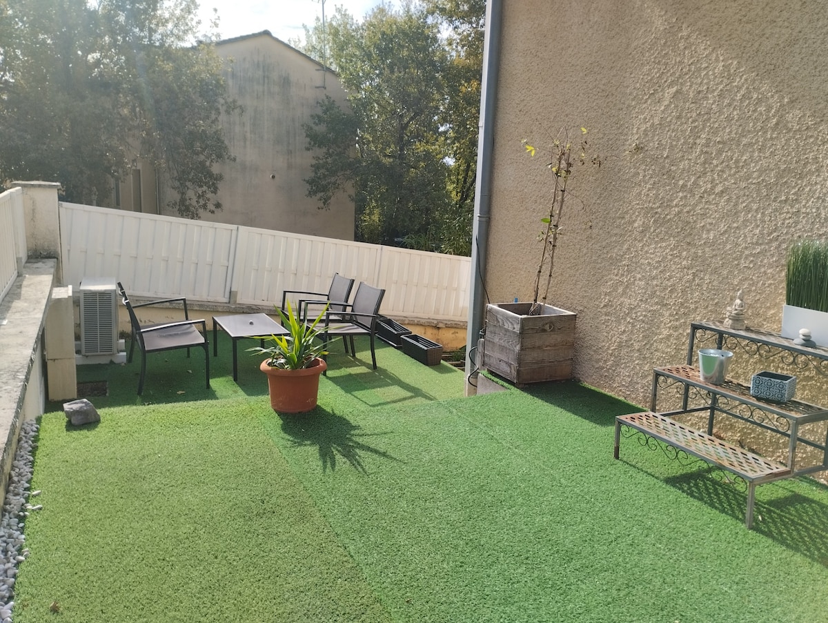 An outdoor area is presented, featuring artificial grass and potted plants. A seating arrangement includes four chairs and a small table, providing a comfortable space for relaxation. An additional planter and a decorative shelf complete the area, enhancing its appeal for outdoor gatherings.