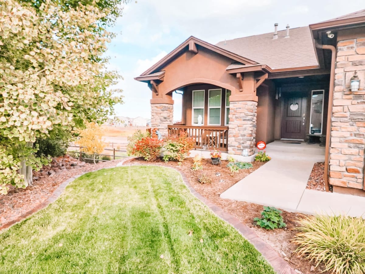 The image captures the entrance of a home, featuring a stone exterior and a wooden porch. A well-manicured lawn frames the walkway, accentuated by greenery and flower beds. The porch is partially shaded by overhanging eaves, creating a welcoming approach to the entrance.