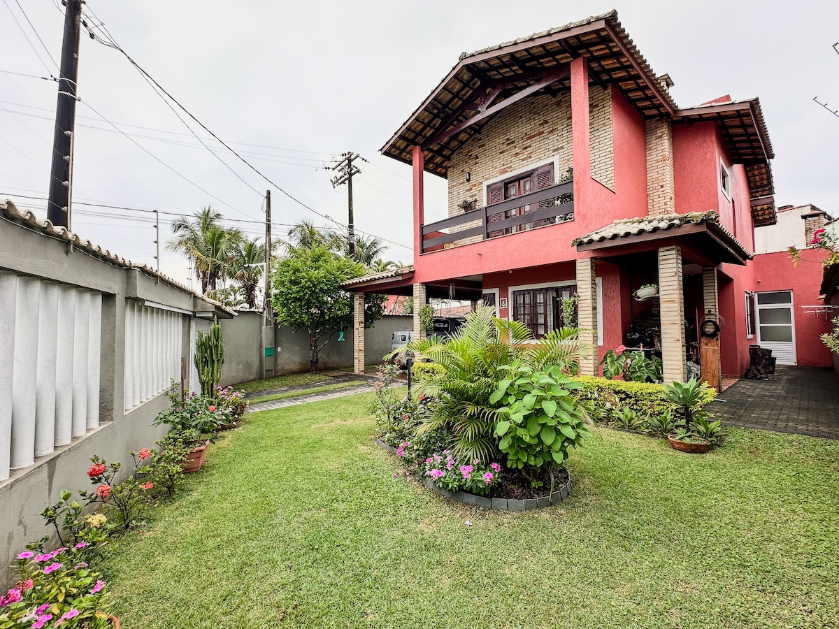 The exterior view of a two-story red house is shown, surrounded by a lush garden. Spacious green lawn is adorned with colorful flowers and bordered by low stone walls. A pathway leads to the entrance, while vehicles are visible under the covered area beside the house.