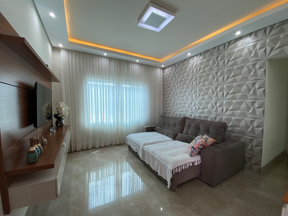 A spacious living area features a modern gray sectional sofa, adorned with colorful cushions. Large windows are dressed with sheer white curtains, allowing natural light to fill the room. A sleek media unit supports a flat-screen television, set against a textured wall with a geometric pattern.