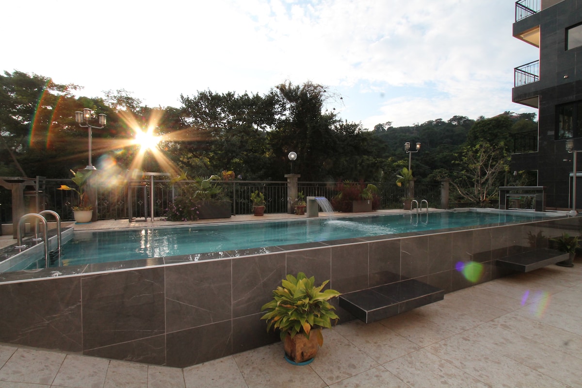A tranquil swimming pool is set against a backdrop of greenery, with gentle sunlight reflecting off the water. Potted plants are placed around the pool area, and steps lead from the pool to the surrounding patio, providing easy access.