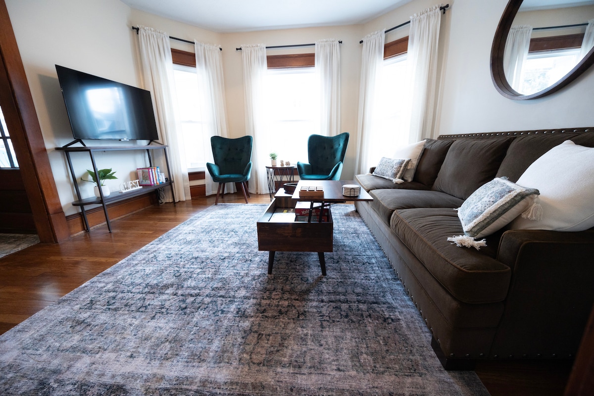 A comfortable living area features a dark sofa paired with two green accent chairs. A wooden coffee table sits in the center, and a television is positioned on the wall opposite. Natural light filters through the large windows, accented by light curtains.