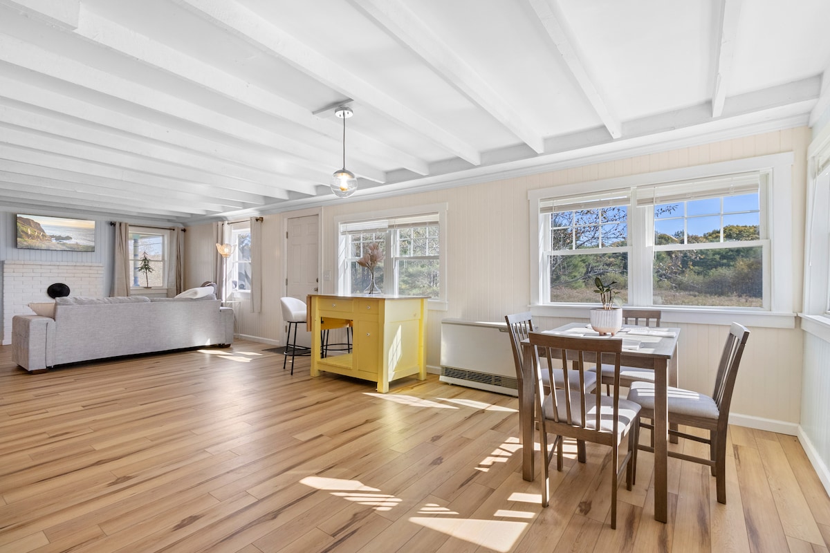 A spacious living area features light wood flooring and exposed beams. A comfortable couch sits adjacent to large windows allowing natural light. A bright yellow island with seating is positioned near a dining area with a table and chairs, creating an inviting space.