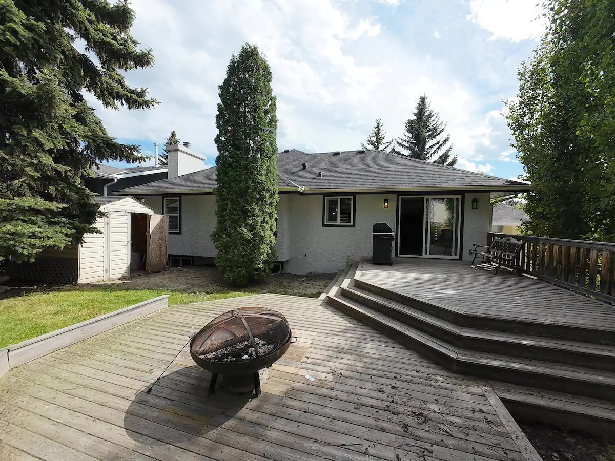 An outdoor deck area features a wooden platform with steps leading down to a grassy lawn. A fire pit is positioned on the ground, surrounded by wooden seating. Lush trees and shrubs frame the backyard, while a sliding glass door reveals access to the interior space.