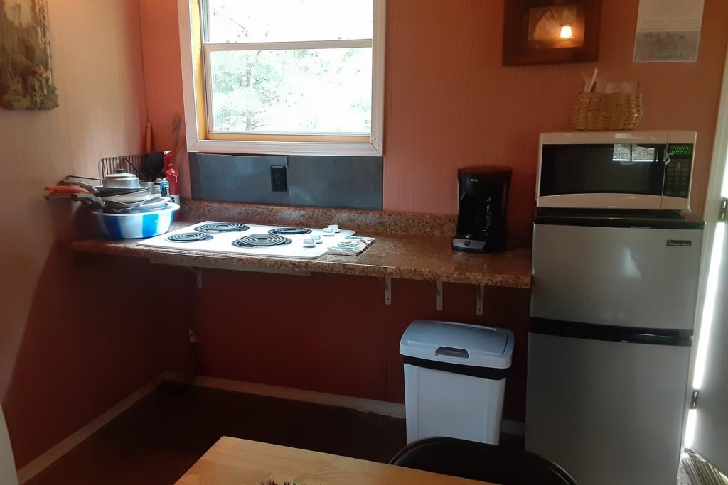 The small kitchen is equipped with a refrigerator, a microwave, and a coffee maker. A countertop with four electric burners is visible, accompanied by various kitchen utensils and dishware. Natural light enters through a nearby window, illuminating the space.