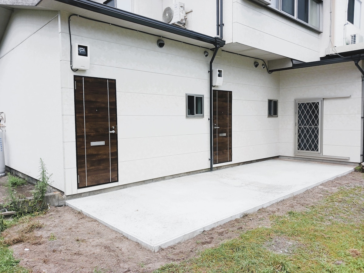 The exterior view captures a clean and modern building with two doors leading to individual rooms. A small window is visible beside each door. A concrete patio area is present, surrounded by a grassy landscape, providing a private outdoor space.