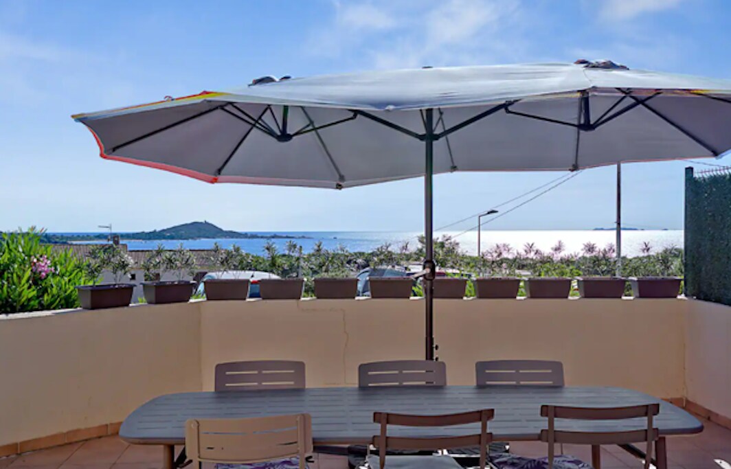 A spacious terrace features a large dining table complemented by several chairs. An expansive umbrella provides shade, overlooking a view of the sea and surrounding greenery. Flower pots adorn the terrace edges, creating a relaxed outdoor dining atmosphere.