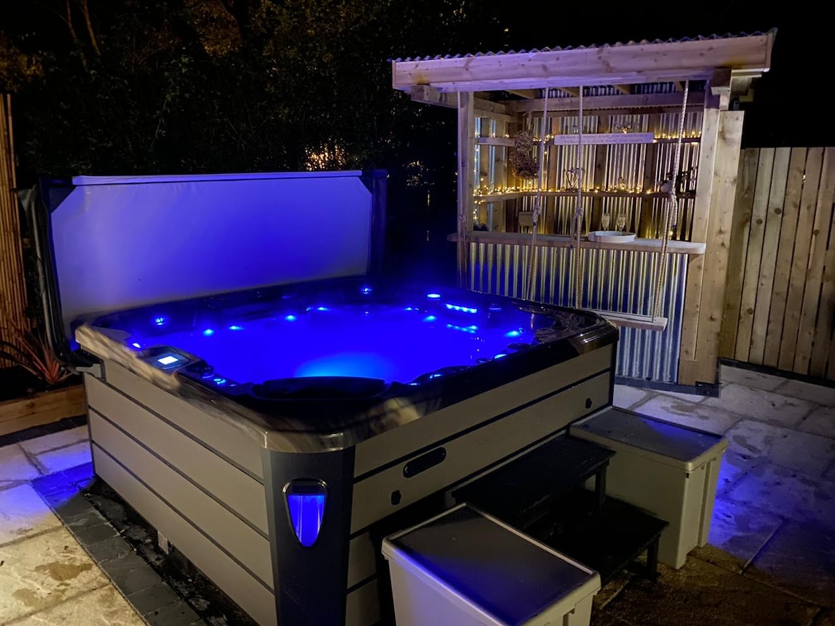 A hot tub spa is illuminated with blue lights, nestled in a private corner of the garden. A wooden shelter with a slatted design stands nearby, offering a shaded area. The surrounding area is composed of paved stones, providing a tranquil setting.