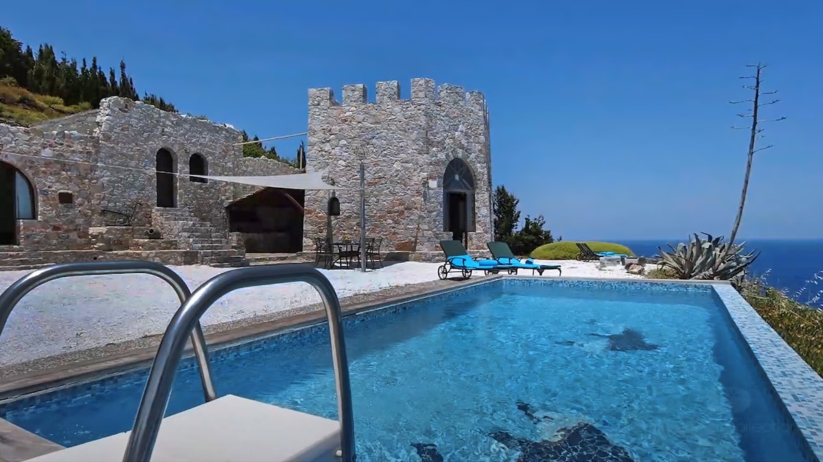 A stone-crafted tower stands prominently against a clear blue sky, surrounded by lush greenery. A sparkling pool reflects the sunlight, framed by lounge chairs positioned for relaxation. The expansive landscape includes distant views of the sea, enhancing the tranquil outdoor setting.