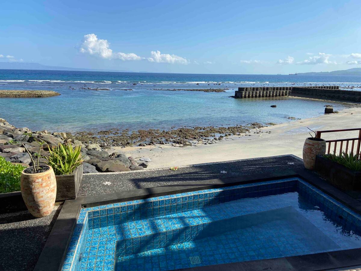 A private pool is situated at the edge of the property, offering views of the tranquil ocean scene. The calm waters, with patches of rock and sandy beach, stretch out towards the horizon. Potted plants frame the pool area, creating a relaxing outdoor space.