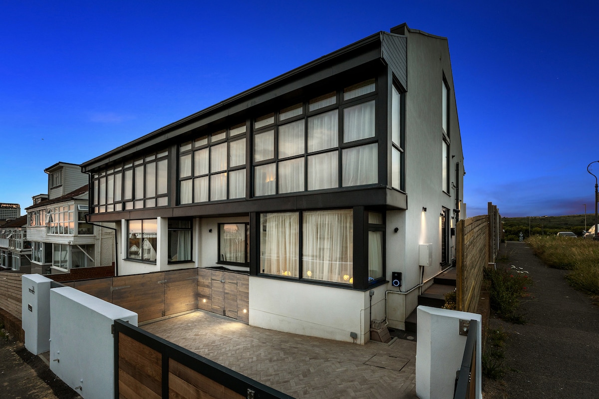 The exterior of a modern semi-detached house is captured at dusk. Large windows dominate the façade, allowing soft light to emanate from within. A spacious driveway leads to the entrance, framed by a wooden fence and grassy area, creating a welcoming first impression.