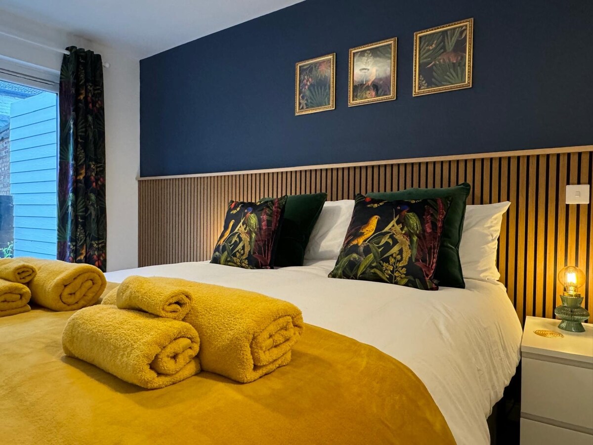 A serene bedroom features a spacious bed dressed in crisp white linens, complemented by vibrant yellow blankets and towels. Framed artwork with tropical themes adds interest to the dark blue wall, while natural light enters through a nearby window, enhancing the calming atmosphere.