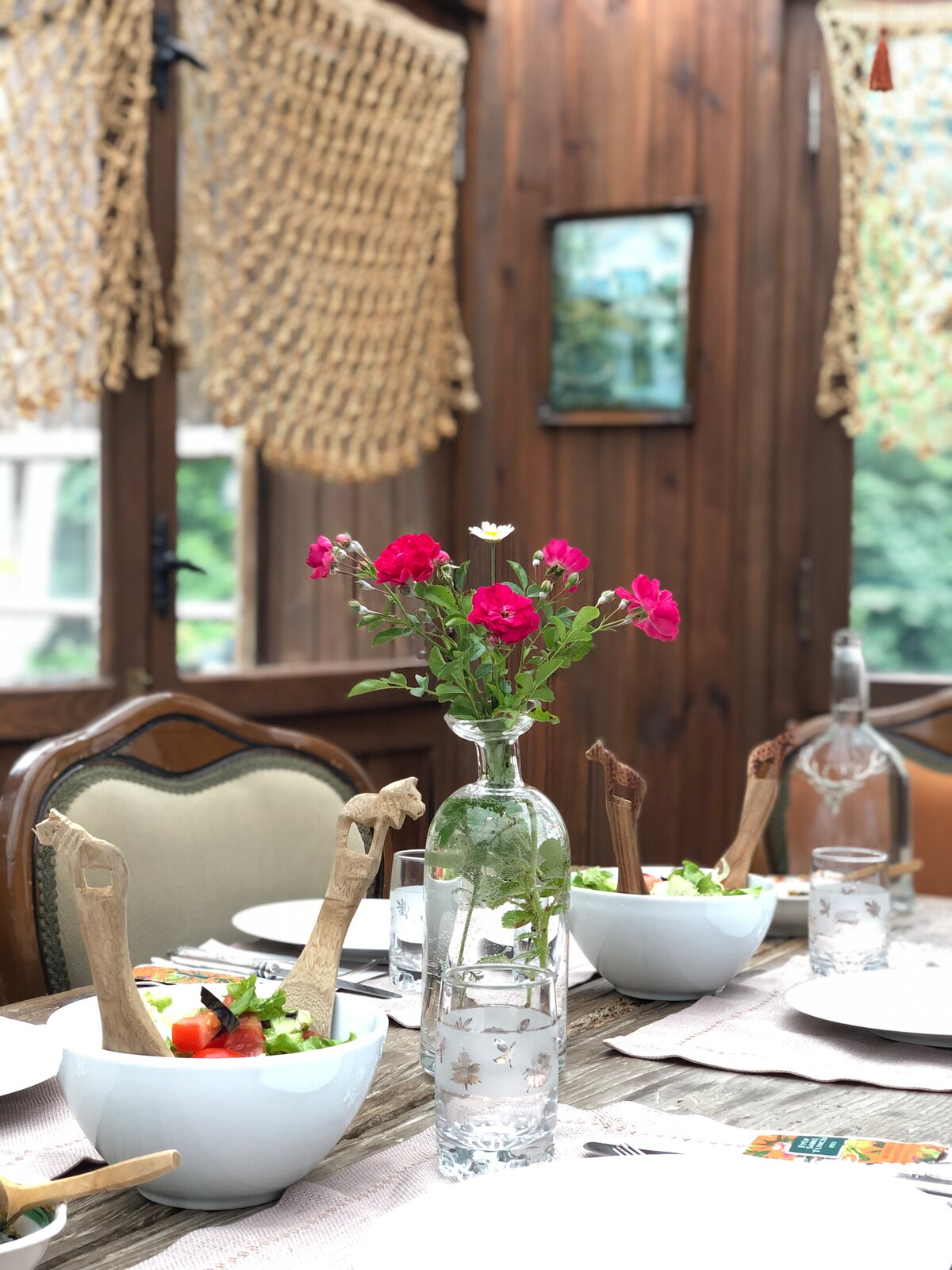 A dining table is set with white bowls containing a variety of salads. A glass vase holds fresh pink flowers, adding a touch of color. Wooden accents and woven decor complement the warm wood paneling surrounding the cozy space.