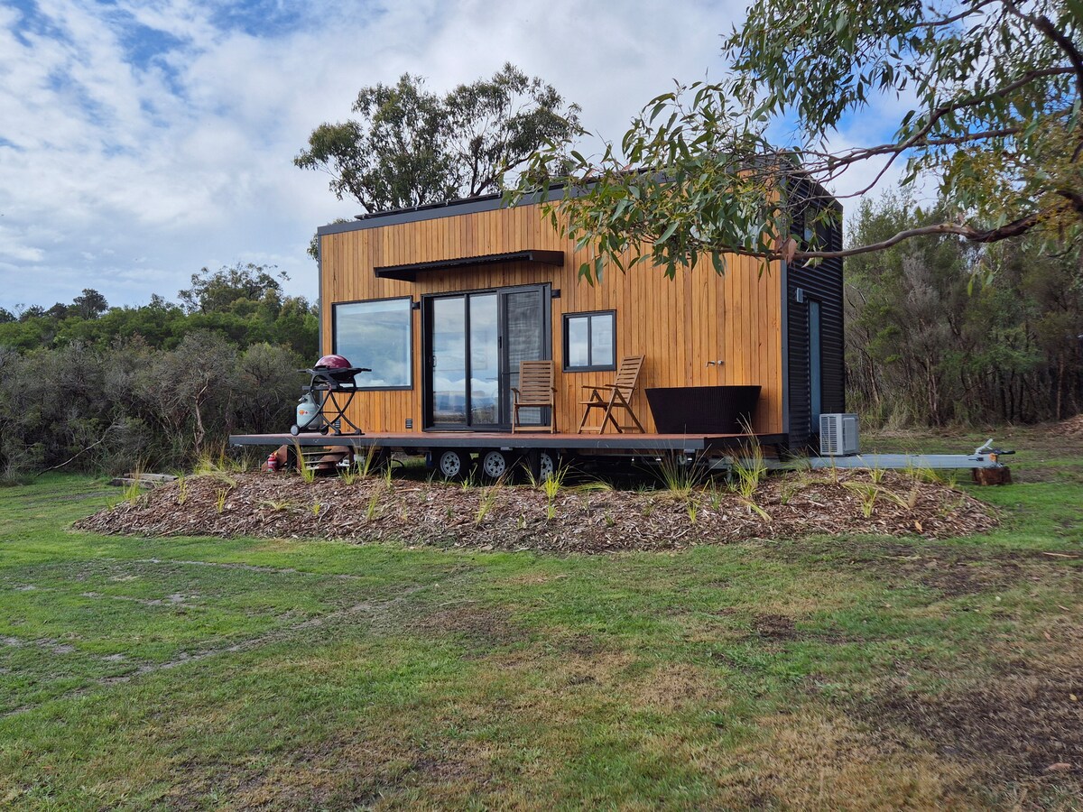 A modern tiny house with wooden cladding is set on a landscaped area, surrounded by native vegetation. Large glass sliding doors offer views of the outdoors. A small deck hosts two wooden chairs and an outdoor BBQ, inviting relaxation in the serene environment.