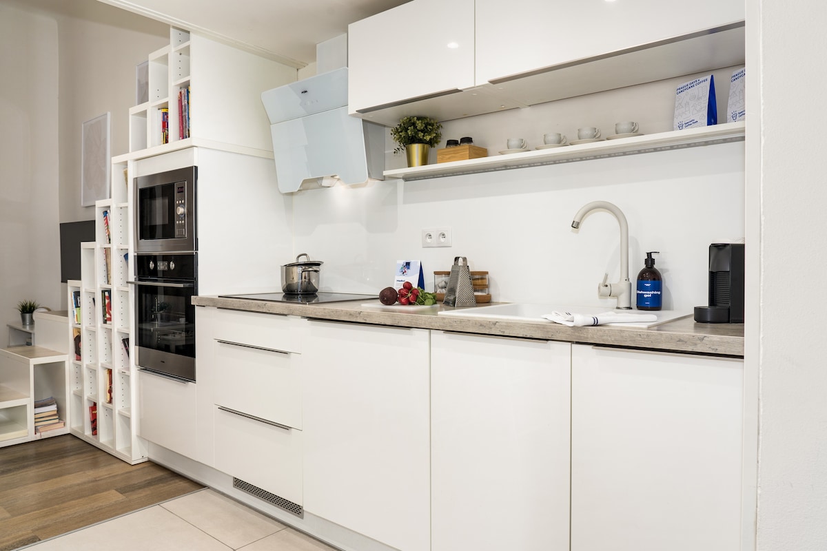 A modern kitchen featuring sleek white cabinetry and a clean countertop. Kitchen appliances include an induction hob, oven, and kettle. Specialty coffee and tea are provided, alongside a compact dining table and a shelf displaying various utensils and items for convenience.