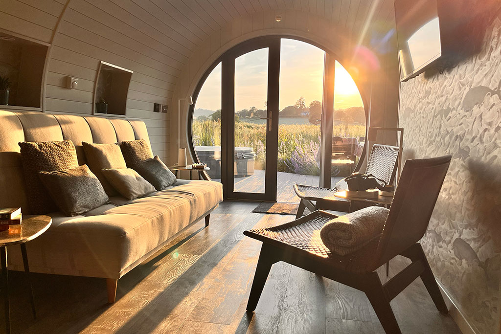 A modern living area features a comfortable sofa with plush cushions, positioned near large glass doors that frame a sunset view. A cozy armchair sits to the side, and natural light illuminates the wood floor, creating a warm atmosphere.
