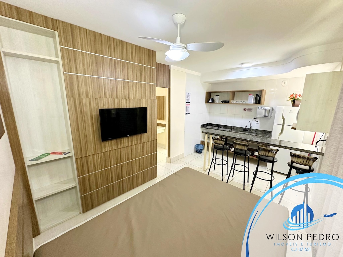 A spacious room features a large bed with a light-colored blanket, a flat-screen TV mounted on a wooden wall panel, and a ceiling fan. A kitchenette is visible in the background, equipped with bar stools and counter space.
