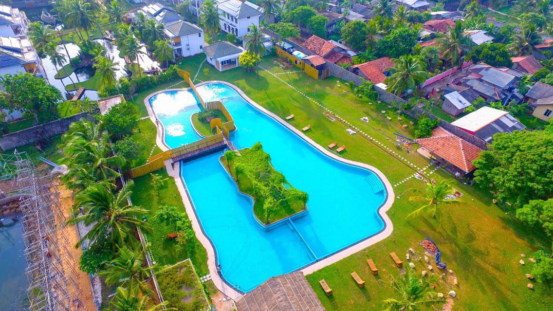 An aerial view displays a large, curvy pool surrounded by lush greenery and seating areas. A central island in the pool is bordered by wooden decking. Lush lawns and tropical trees create a serene atmosphere, with neighboring structures visible in the background.