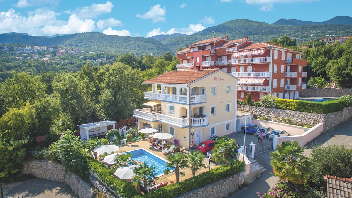 A spacious villa is set amid lush greenery, featuring a central pool surrounded by lounge areas with umbrellas. Multiple levels of the villa provide a harmonious blend with the natural surroundings. Scenic mountains rise in the background, showcasing the peaceful hillside location.