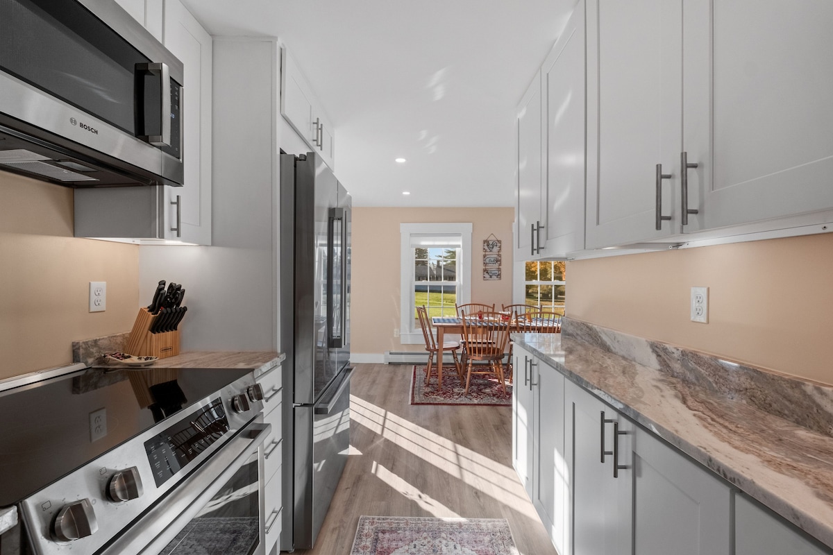 A modern kitchen is presented with light cabinets and expansive countertops. Stainless steel appliances, including a range and refrigerator, are visible. Natural light fills the space, illuminating the dining area with a wooden table and chairs in the background, framed by a large window.