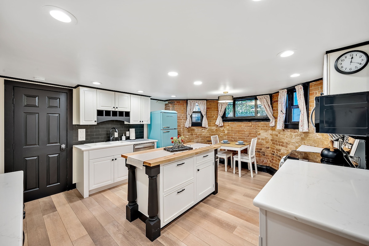 The spacious kitchen features light cabinetry complemented by a central island with a wooden countertop. Exposed brick walls provide a historic touch. Large windows allow natural light, while a dining table with chairs is positioned nearby, creating a functional and inviting cooking space.