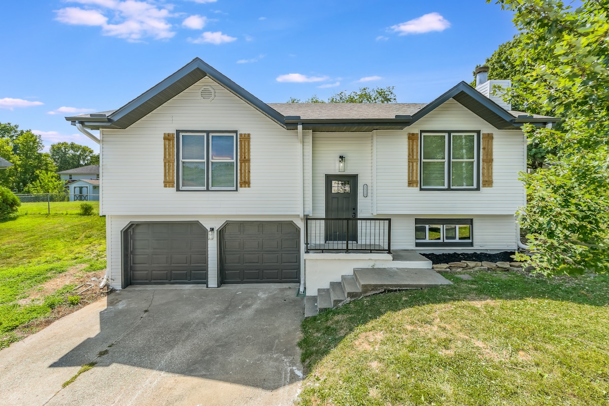 The exterior of the home features a light-colored facade with wooden accents around the windows. A neatly manicured lawn and driveway lead to a two-car garage below the main entrance, framed by greenery in the background under a clear blue sky.