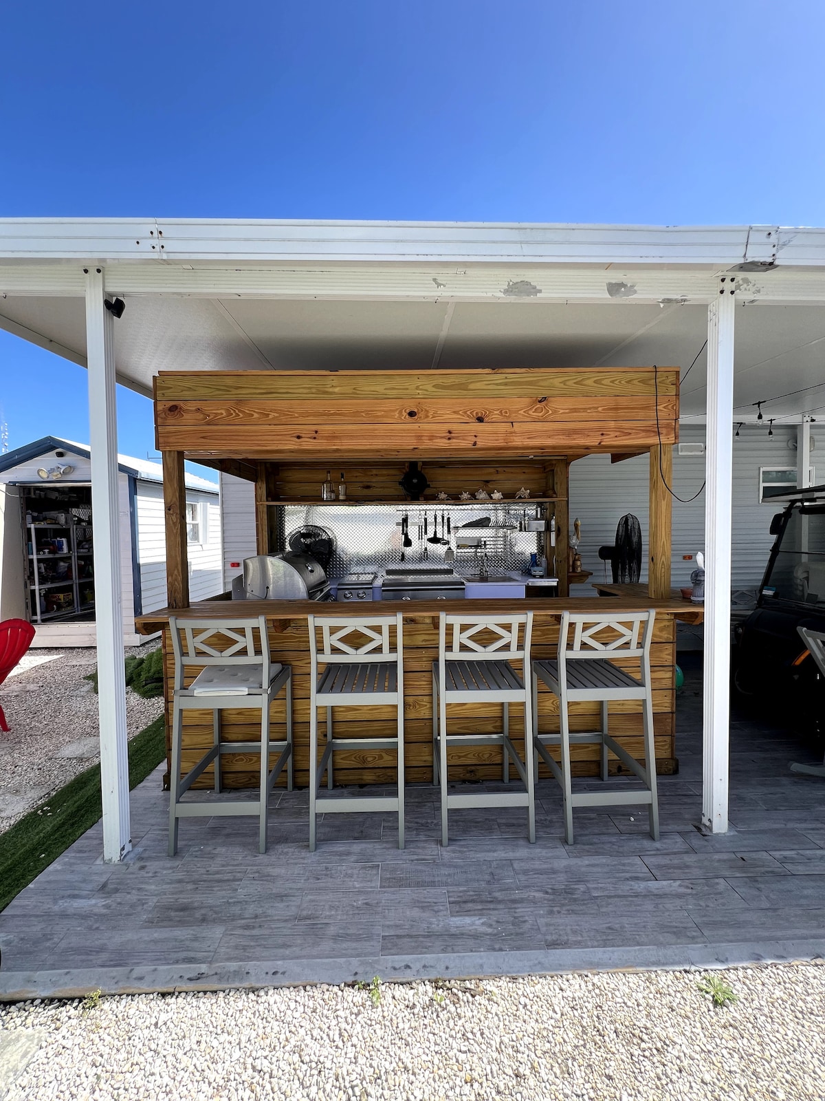 An outdoor hangout bar features a wooden countertop with high stools arranged in front. The space is well-lit, allowing for both cooking and socializing. The backdrop includes a glimpse of nearby mobile homes, and a clear blue sky enhances the inviting atmosphere.